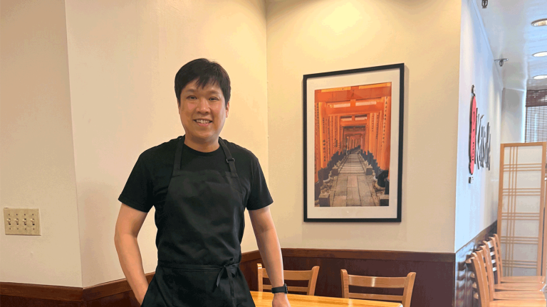 Man in black apron standing in front of a picture inside a Japanese restaurant.
