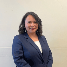 A woman in a blue suit jacket and shoulder-length black hair smiling in front of a white background.