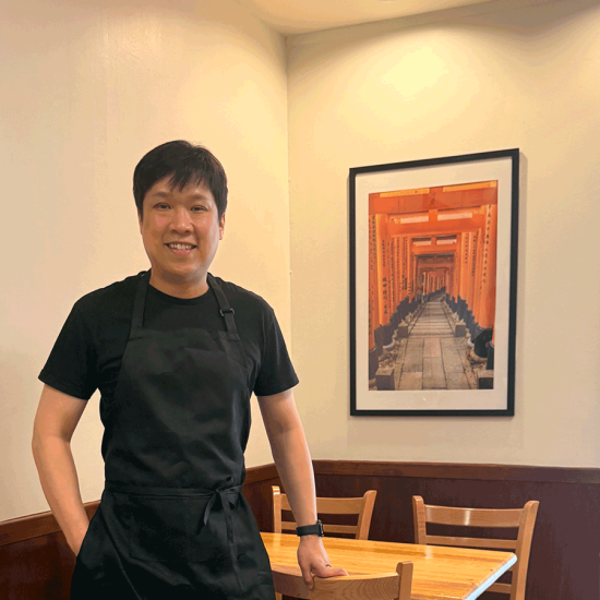 Man in black apron standing in front of a picture inside a Japanese restaurant.
